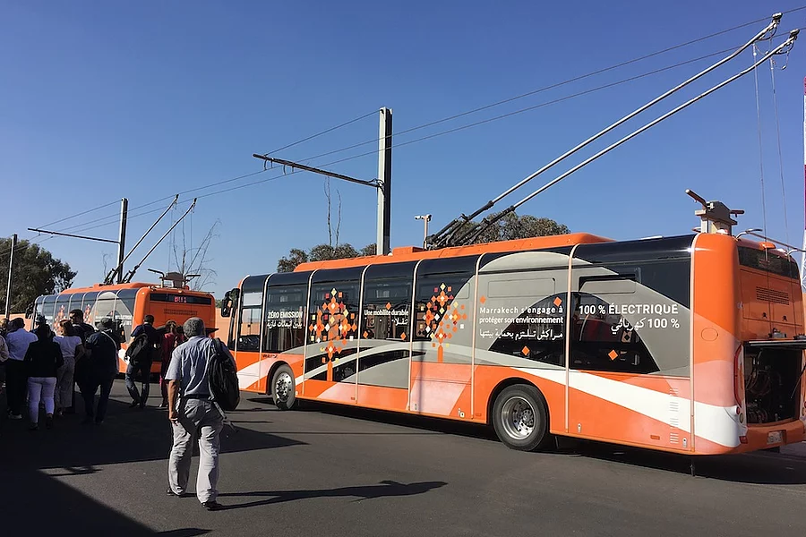 L'image montre un bus électrique orange à Marrakech, au Maroc, arborant un design moderne et des slogans tels que « 100 % électrique » pour promouvoir des transports en commun respectueux de l'environnement.