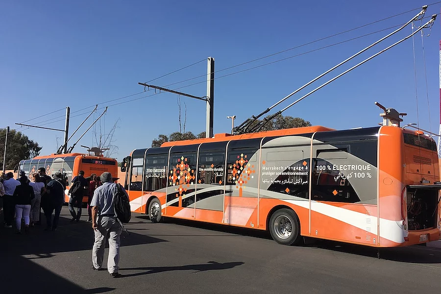 L'image montre un bus électrique orange à Marrakech, au Maroc, arborant un design moderne et des slogans tels que « 100 % électrique » pour promouvoir des transports en commun respectueux de l'environnement.