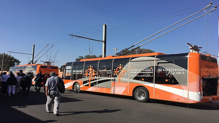 L'image montre un bus électrique orange à Marrakech, au Maroc, arborant un design moderne et des slogans tels que « 100 % électrique » pour promouvoir des transports en commun respectueux de l'environnement.